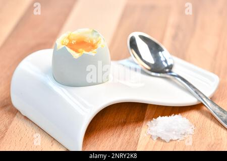 a single boiled egg in an egg cup broken open showing yolk with a spoon on a wooden surface Stock Photo