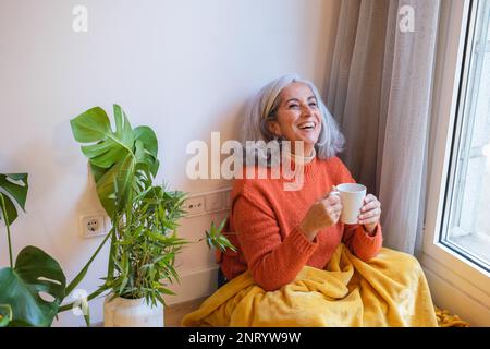 Senior woman with gray hair drinking herbal tea sitting on the floor and relaxing at home. Concept: Lifestyle, senior life, relaxation Stock Photo