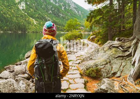 Young blond hair woman with big camping rucksack wearing yellow raincoat jacket, purple and blue cap looking on mountains trail near lake. Copy space, Stock Photo