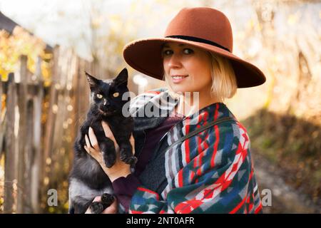 Black cat and stylish traveling woman wearing  authentic boho chic style poncho and hat near old wooden countryside buildings Woman have a fun in autu Stock Photo