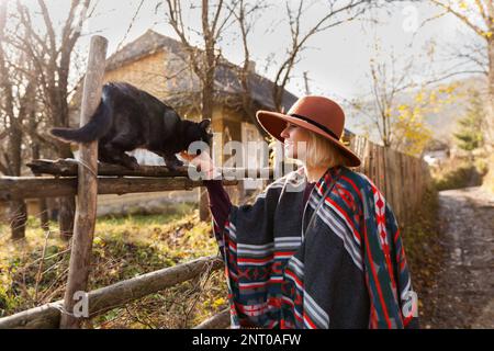 Black cat and stylish traveling woman wearing  authentic boho chic style poncho and hat near old wooden countryside buildings Woman have a fun in autu Stock Photo