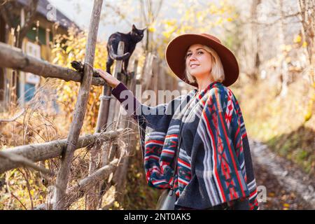 Black cat and stylish traveling woman wearing  authentic boho chic style poncho and hat near old wooden countryside buildings Woman have a fun in autu Stock Photo