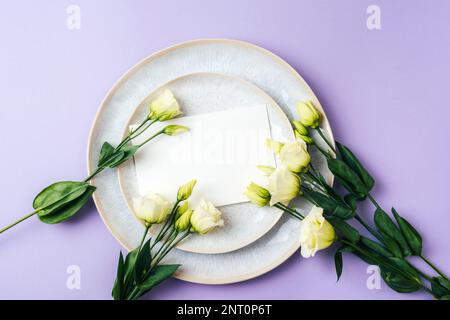 Eustoma grandiflorum white flowers and blank card on the plate on purple background, mockup. Festive background, flat lay, copy space. Stock Photo