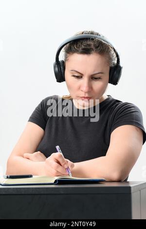 Busy young woman in headphones working on laptop, making video call and making notes in notebook, work and study online, copy space and white backgrou Stock Photo