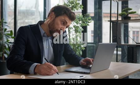 Busy successful caucasian adult businessman in office typing on laptop talking to client on mobile phone writing notes explain condition multitasking Stock Photo