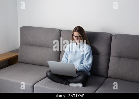 Young white woman sitting on sofa at home and working on laptop. Beautiful blonde female typing text on laptop on couch in the living room Stock Photo
