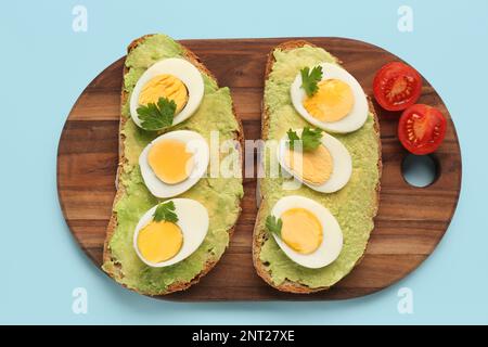 Board with delicious avocado toasts, boiled eggs, parsley and tomatoes on blue background Stock Photo