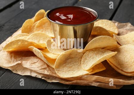 Parchment with delicious potato chips and bowl of ketchup on black wooden background Stock Photo