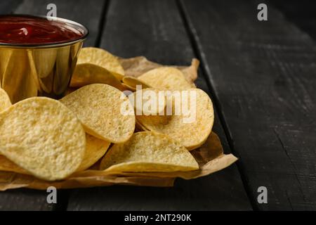 Parchment with delicious potato chips and bowl of ketchup on black wooden background Stock Photo
