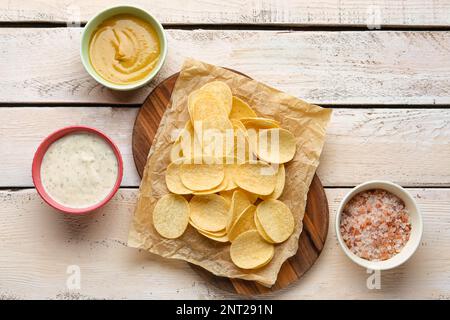Board with delicious potato chips, sauces and sea salt on white wooden background Stock Photo