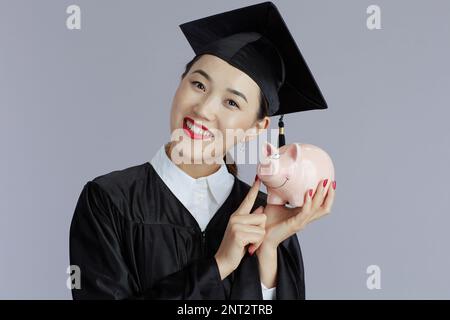 smiling modern female asian graduate student with piggy bank against gray background. Stock Photo