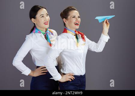 smiling elegant stewardess women in blue skirt, white shirt and scarf with paper airplane against gray background. Stock Photo