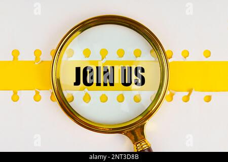 Between two sheets from a notebook on a yellow strip with the inscription - join us, there is a magnifying glass. Business concept. Stock Photo