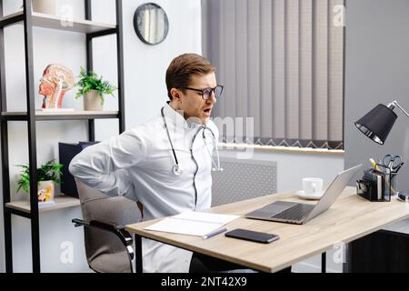 A young male doctor sits at his workplace and feels pain in his back. A doctor takes a break from his work day because of pain. Stock Photo