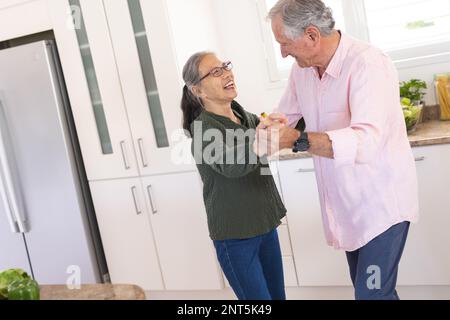 Happy senior diverse couple dancing together in kitchen Stock Photo