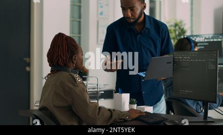 Software developer holding laptop and talking to programer at desk, working on app development and running code. System engineers coding and writing machine learning database. Stock Photo