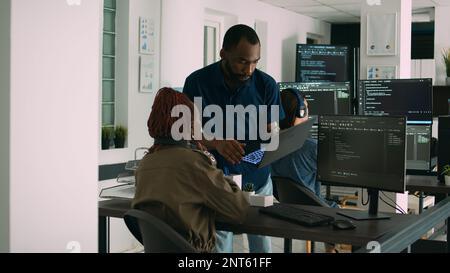 Two software coders analyzing source code looking for errors on laptop, checking big tv monitor in it agency office. Programmers working on cyber security innovation in busy company space. Stock Photo