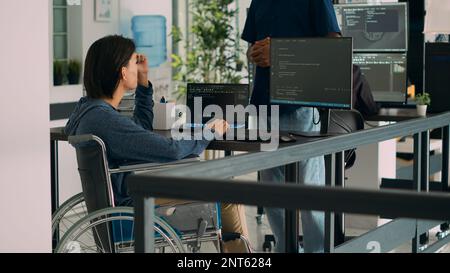 App developer talking to coder with chronic impairment in agency office, explaining source code programming on computer. Wheelchair user coding script with new user interface. Stock Photo