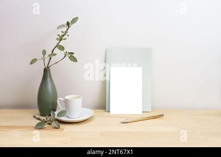 Blank greeting card or invitation mock-up with a pen next to a gray green vase with sage branches and a coffee cup, simple home decor style, copy spac Stock Photo