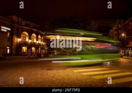 A green city bus drives down a night street in the light of street lamps past the Landskrona Theatre. Long exposure. Blurred lights of a traveling tar Stock Photo