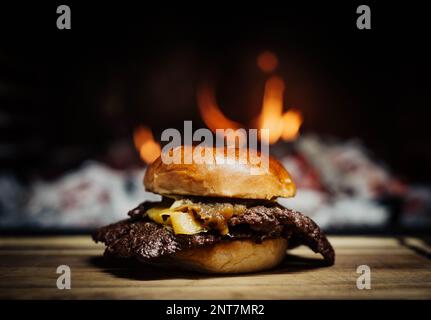 Smashed Cheeseburger on a wooden table in front of a fireplace. Stock Photo