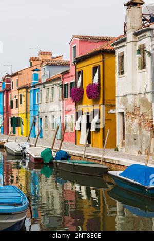 Moored boats on canal lined with colourful stucco houses decorated with striped curtains over entrance doors and windows, Burano Island, Italy. Stock Photo