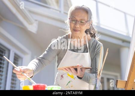 Happy senior asian woman painting on easel in garden Stock Photo