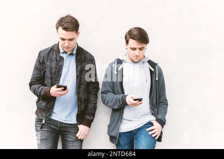 two antisocial mobile phone addicted male teenagers looking at their smartphone, technology concept, leaning against wall with copy space Stock Photo