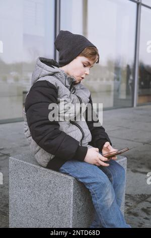 7 year old boy playing with smartphone, sitting outside in winter jacket and knit hat Stock Photo