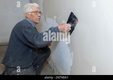 an old man holding wood trowel is plastering cement wall at home, concept a hobby in elderly people.pensioner worker plastering the wall.senior worker Stock Photo