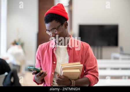 Satisfied hipster black student guy stands in college library with textbooks using phone break study Stock Photo