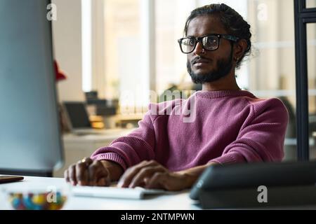 Serious young programmer in eyeglasses working online on computer at his workplace in IT office Stock Photo