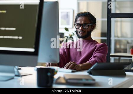 Young developer in eyeglasses concentrating on his work on computer sitting at table in IT office Stock Photo