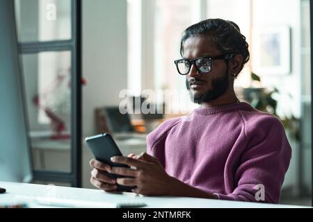 Young programmer in eyeglasses using mobile phone in his online work at table Stock Photo