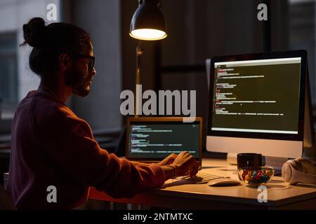 Young programmer sitting at his workplace and working on computer with codes, he working in office till late night Stock Photo