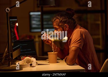 Young programmer examining codes on computer monitor and having lunch while working till late night in office Stock Photo
