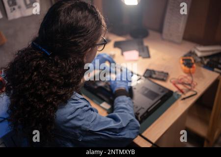 Technician repairing a laptop in the old lab. Young woman working, wearing gloves in retro workshop. Concept of repair computer, electronic, upgrade, Stock Photo