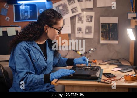 Female technician repairing broken laptop notebook computer with a screwdriver in retro repair shop. Copy space Stock Photo