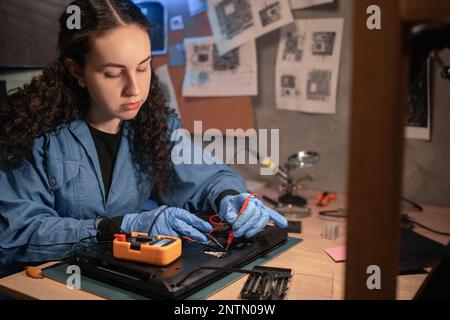 Concentrated female technician busy repairing laptop and it testing with multimeter at retro workshop. Concept of professional worker and repair servi Stock Photo