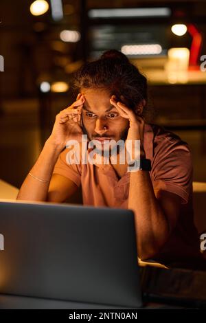 Vertical image of young programmer looking very tired sitting at his workplace, he working over computer program in dark office Stock Photo