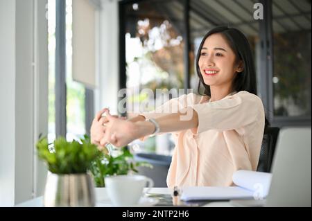 Beautiful and smiling millennial Asian businesswoman stretching her arms at her desk, feeling happy after finished work, resting in the office. Stock Photo