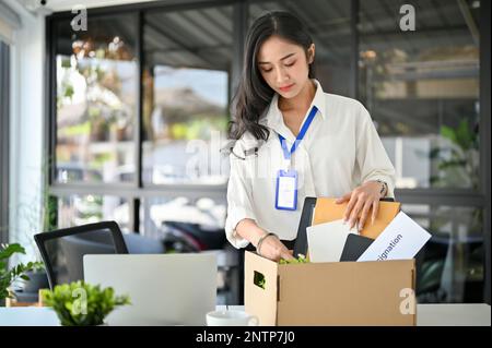 Sad and unhappy millennial Asian female office worker packing her stuff into a cardboard box in the office, quitting her job or getting fired from her Stock Photo