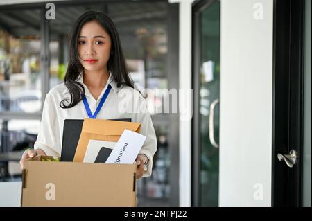 Sad and unhappy young Asian female office worker carrying a cardboard box with her belongings and resignation letter, being fired from her boss, walki Stock Photo