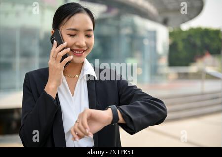 Successful and professional millennial Asian businesswoman or female CEO is on the phone with business partner and checking the time on her wristwatch Stock Photo