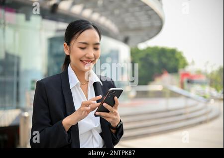 Attractive and beautiful millennial Asian businesswoman using her phone, checking email while walking outside along the company building. urban busine Stock Photo