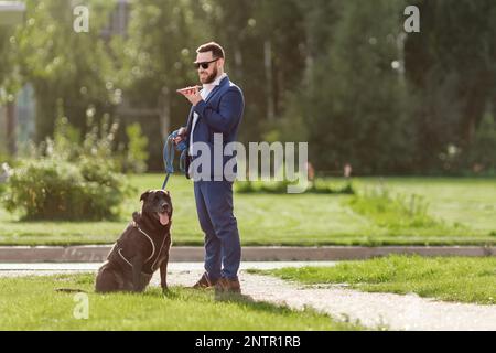 Businessman in stylish suit talking on mobile phone and holding his dog sitting on leash on urban park background Stock Photo