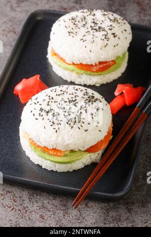 Sushi menu with burger made from rice and smoked salmon, avocado, black sesame and ginger close-up on a plate on the table. Vertical Stock Photo