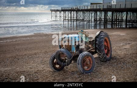 A vintage and rusted tractor used by a fisherman to bring in his boat, lies parked on the shingle beach in front of Bognor Regis Pier, in West Sussex, Stock Photo