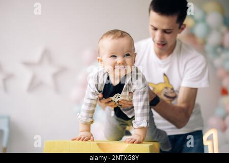 Happy father plays with his son celebrating first birthday of baby boy against background of colorful balloons. Celebration first birthday. Festive ev Stock Photo