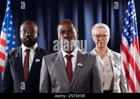 Group of confident intercultural male and female politicians in formalwear looking at camera while standing between two American flags Stock Photo